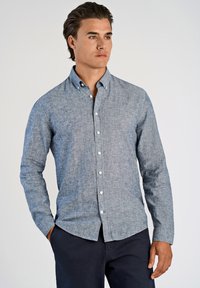 Camisa de botones de manga larga en tejido de espiga azul claro. Presenta un cuello clásico y botones en la parte delantera. Combinada con pantalones oscuros.