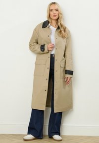 Cappotto lungo beige con collo e polsini neri, dotato di grandi tasche frontali e chiusure a scatto. Tessuto liscio con una vestibilità comoda.