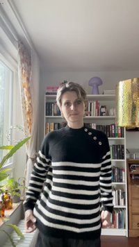 Mustriline mustvalge sviiter kõrge kaelusega, millel on kolm suurt hõbedast nööpi õlal ja pehme, tekstuuriga pind.