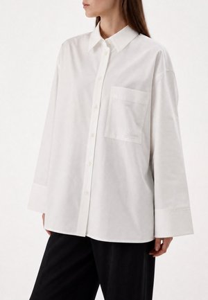 Femme portant une chemise blanche ample à boutons avec une poche poitrine et un pantalon noir, montrée de la moitié du torse au cou et aux hanches.