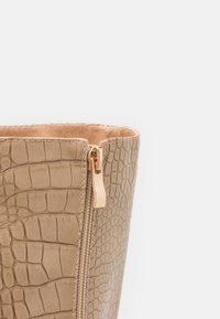 Sac en similicuir beige à texture crocodile, doté d'une fermeture éclair dorée. L'intérieur est doublé d'un matériau léger et lisse.