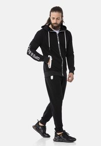 Schwarze Zip-Hoodie mit weißen Akzenten, Fronttaschen und Logo am Ärmel; kombiniert mit schwarzen Jogginghosen und sportlichen schwarzen Sneakern.