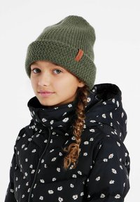 Groene gebreide beanie met een gestructureerd patroon en een bruine leren label. Draagt bij een zwarte gewatteerde jas met een wit bloemenpatroon.