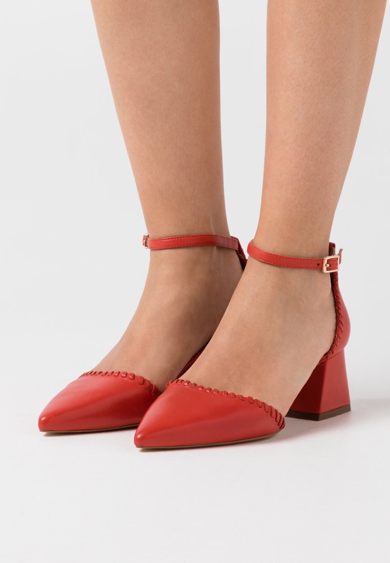 Escarpins en cuir rouge à bout pointu avec une sangle de cheville et un talon carré. Présente des coutures décoratives le long du bord. Texture lisse.