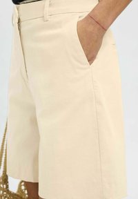 Pantaloni corti beige in cotone, dotati di tasche laterali, taglio dritto e superficie liscia. La vita è rifinita con passanti per cintura.