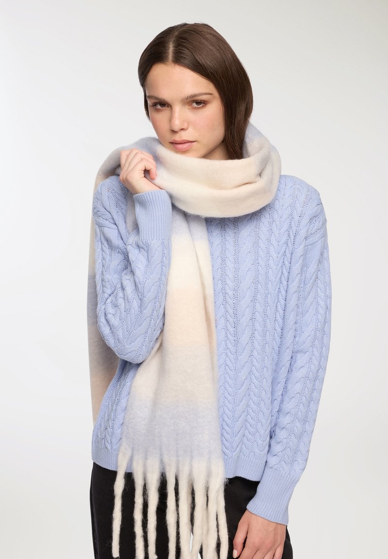 Jasnoniebieski sweter w warkoczowy wzór w parze z długim, miękkim szalem w odcieniach kremowym i jasnoniebieskim, z frędzlami na krawędziach. Gładka tekstura, ciepłe materiały.