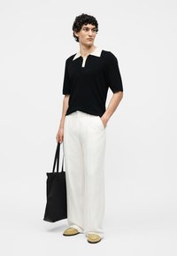Homme portant un polo noir avec col blanc, pantalon blanc à jambes larges, chaussures jaunes, tenant un sac fourre-tout noir, debout sur fond blanc.