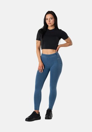 Sinivihreät leggingsit, joissa on korkea vyötärö ja joustavasta kankaasta valmistetut. Yhdistetty mustaan lyhytpaitaiseen t-paitaan ja mustiin urheilukenkiin. Yksinkertainen muotoilu.