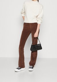 Pantalon évasé en velours côtelé marron, pull en tricot crème avec ourlet côtelé, sac à main noir avec fermoir et bandoulière chaîne, baskets blanches avec lacets noirs.