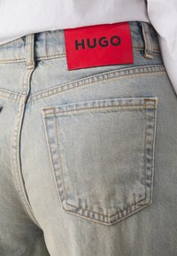 Světle modré denimové džíny s červenou koženou nášivkou s nápisem "HUGO" černým písmem. Standardní zadní kapsa se viditelnými detaily prošívání.