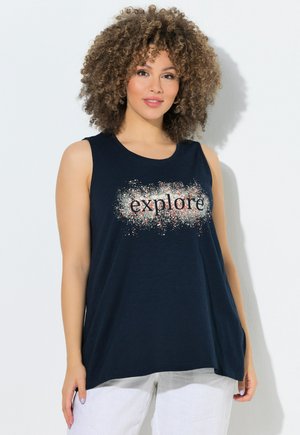 Frau mit lockigem Haar trägt ein ärmelloses schwarzes Oberteil mit dem Schriftzug „explore“ in weißer und oranger Klecks-Optik, kombiniert mit weißen Hosen.