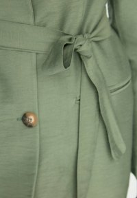 Vihreä voitokas takki, jossa on sidottava vyö, teksturoitu kangas ja messinkinen nappi. Sisältää pienen taskudetaalin sivussa.