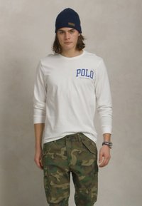 Λευκό πουκάμισο με μακριά μανίκια και μπλε κείμενο "POLO", συνδυασμένο με πράσινες παραλλαγές cargo παντελόνες. Το μοντέλο φοράει μια ναυτική μαύρη σκούφια και ένα ρολόι.