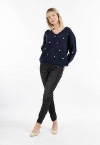 Navy Pullover mit V-Ausschnitt und goldenen Herzmustern, langen Ärmeln, kombiniert mit schwarzen Skinny-Jeans und beigen High Heels. Weiche Stricktextur.