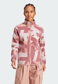 Chaqueta de forro polar en rosa con patrones abstractos, cuello alto, cremallera completa y logo en el pecho. Llevada sobre pantalones cargo beige.