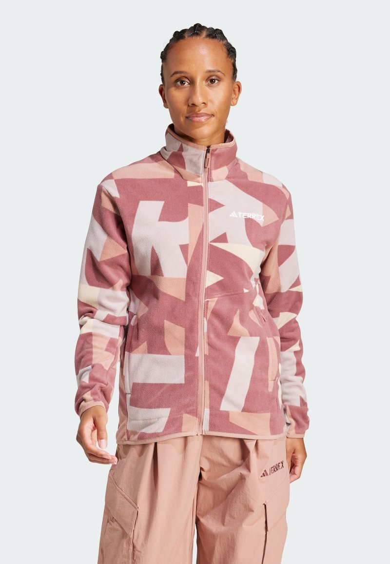 Fleecejacka i rosa med abstrakta mönster, hög krage, hel dragkedja och logotyp på bröstet. Bärs överbeige cargobyxor.