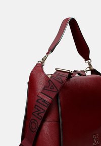 Rode leren handtas met een gestructureerde afwerking, voorzien van een afneembare schouderriem met zwarte opdruk en accenten in goudkleurig metaal.