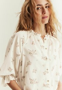 Hvid bluse lavet af tekstureret stof med et blomstermønster i bløde nuancer af lyserød, med rynket halsudskæring og knaplukning foran.