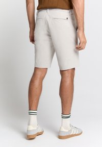 Lichte beige heren shorts van gladde stof, met achterzakken en een klein merkteken, gecombineerd met grijze sneakers en gestreepte sokken.