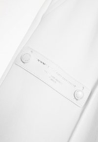 Tela de algodón blanca con superficie texturizada. Presenta una etiqueta blanca de marca con texto en relieve y un botón a presión plateado.