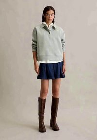 Jovem mulher em pé, vestindo uma polo de manga longa cinza, uma camisa branca, uma saia plissada azul-marinho e botas de couro castanhas até o joelho, em um fundo neutro.