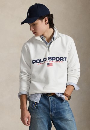 Biała bluza polarowa z zamkiem do połowy długości i napisem "POLO SPORT" w kolorze granatowym i czerwonym, nałożona na jasnoniebieską koszulę w paski. Jeansy denimowe i niebieska czapka.