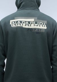 Donkergroen hoodie van een gestructureerde stof. Heeft een groot off-white logo op de achterkant met rasterpatronen en geometrische accenten.