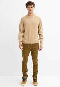 Suéter de punto beige con detalles acanalados, combinado con pantalones cargo verde oliva que cuentan con bolsillos y puños elásticos, además de zapatillas altas marrones.