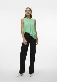 Top sans manches vert menthe avec un décolleté en V, associé à un pantalon large noir. Le modèle porte des chaussures à talons pointues blanches et un collier en argent.