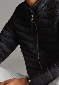 Doudoune noire matelassée avec un col haut, dotée d'une fermeture éclair argentée et d'un tissu texturé. Le bras présente un détail de bouton près du poignet.