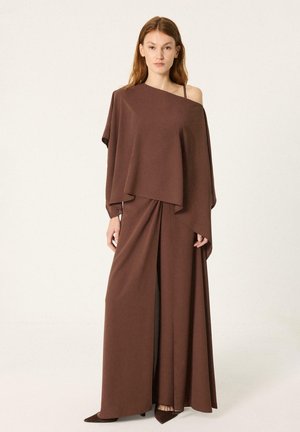 Femme portant un haut marron asymétrique drapé, épaules dénudées, et une longue jupe assortie avec une fente devant, debout sur un fond uni.