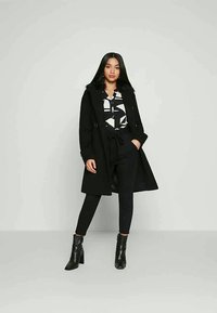 Black wool coat with a fur collar, worn over a white and black patterned shirt, paired with black trousers and ankle boots.