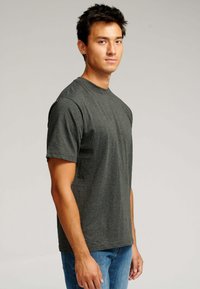 Ung mand står sidelæns, iført en mørkegrå t-shirt med korte ærmer og blå jeans, mod en ensfarvet lys baggrund.
