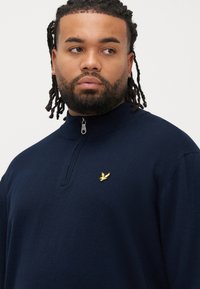 Maglione blu navy con zip al collo realizzato in tessuto morbido, caratterizzato da un logo ricamato in oro sul petto e un colletto a coste.