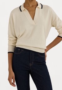 Beige pulovr s hlubokým véčkovým výstřihem a tmavě modrým lemováním u límce a manžet, spárovaný s tmavě modrými jeansy z denimu s oranžovým prošíváním.