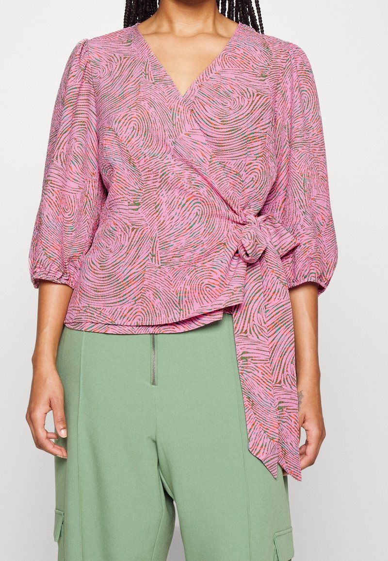 Femme portant un chemisier cache-cœur rose avec des motifs abstraits en spirale et un nœud noué, associé à un pantalon taille haute vert clair avec détail de fermeture éclair.