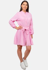 Abito rosa con bottoni, maniche lunghe, tessuto testurizzato, dettaglio con cintura in vita e gonna svasata. Abbinato a sneaker bianche.