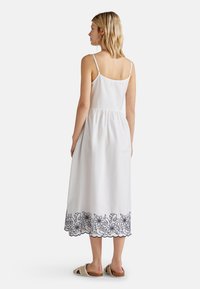 Weißes Baumwollkleid mit Spaghetti-Trägern, geraffter Taille und einem floralem Spitzenrand in Marineblau am Saum. Die Modell trägt lässige Sandalen.
