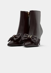 Rudos odinės kulkšninių batų su aštriai formuotu pirštu dizainu, priekyje su dideliais kaspinais ir auksinėmis detalėmis. Vidutinio aukščio kulnas.