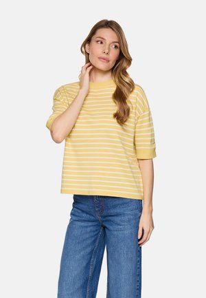 Mujer con cabello largo y ondulado, vestida con una camiseta de manga corta a rayas amarillas y blancas y jeans azules, de pie con la mano cerca del cuello, mirando de lado.