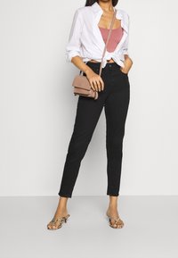 Černé vysoké kalhoty s knoflíkovým zapínáním vpředu, spárované se uzlovanou bílou košilí přes růžové tílko a béžovou crossbody kabelkou. Metalické sandály.