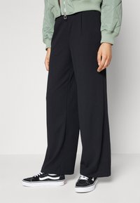 Pantalones de pierna ancha negros con una tela lisa, que presentan pliegues en el frente. Combinados con zapatillas negras y blancas, y una chaqueta bomber acolchada de color verde claro.