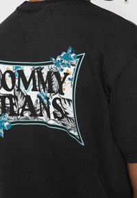 Camiseta negra con un gran gráfico floral en blanco, azul y naranja, con letras en negrita "TOMMY JEANS" en el centro. Diseño de manga corta.