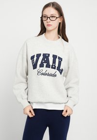 Bluză sport gri deschis cu inscripția „VAIL” în litere bleumarin și „Colorado” scris cursiv dedesubt. Guler rotund, mâneci lungi și manșete rib.
