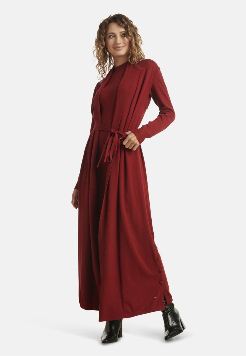 Long red cardigan dress with a belted waist, ribbed texture, side buttons, and long sleeves. Paired with black heeled boots.