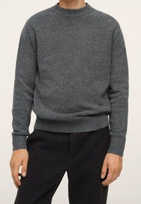 Grauer Strickpullover mit Rundhalsausschnitt, strukturiertem Stoff, langen Ärmeln und geripptem Saum. Kombiniert mit dunkler Hose.