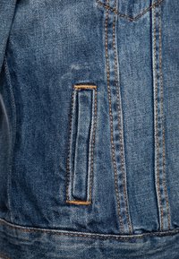 Denimjacka i blå nyans, med synlig sömnad i orange och en smal vertikal ficka på framsidan, som visar en texturerad tyg.