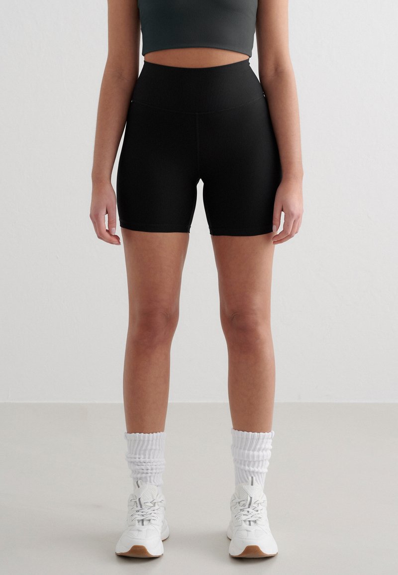 High-waisted black shorts made of ribbed fabric. Features a smooth waistband and a fitted silhouette, paired with white athletic shoes and socks.