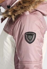 Doudoune avec une section supérieure rose et une section inférieure matelassée blanche, dotée d'une capuche bordée de fourrure et d'un écusson logo noir sur la manche.