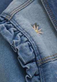 Denim stof in een lichtblauwe tint, met een gerimpelde rand en een kleine bloemembroidery in roze en geel. Zichtbare stikdetails.